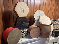 Photo showing various vintage hat boxes stacked against a wood-paneled wall with visible branding of Jordans, Cherry and Company, and Star Store.