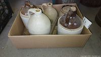 Photo showing four stoneware jugs and a covered crock in a cardboard box; jugs are beige and one has a brown glazed top.