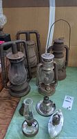 Photo showing four large metal oil lanterns with glass chimneys and metal handles in background, four smaller oil lamps in foreground including porcelain one and metal-based ones.