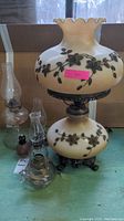 Photo showing the parlor lamp with cream-colored glass and dark metal floral decor, ruffled top, metal base, surrounded by five assorted oil lanterns on a green surface.