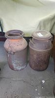 Two rusted metal milk churns side by side on the floor, showing general condition and size.