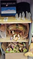 Boxed seven-piece frosted glass nativity set with visible figurines on the shelf above a wooden stable containing colorful resin nativity figures and a box of straw on the side.