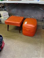 Square orange vinyl padded foot stool with wooden tapered legs and scratches on surface next to oval padded orange ottoman