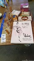 Photo showing all items arranged on wooden table: six novelty pin-up girl drinking glasses, several boxed novelty gag gifts including 'For Milady' Adam to Eve design, 'Little Squirt' surprise toy, 'Pe-cans' candy box, 'Tom & Paul Pot Brothers' box, Henry 8th mug box, and Merry Christmas cards/game box.