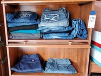 View of assorted women's jeans and pants folded and stacked on two shelves inside a wooden cabinet. Various denim shades and fabrics visible.