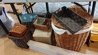 Overview of assorted laundry and wicker baskets in different sizes and styles placed on the floor under a table, showing variety including lined and unlilned baskets, some with lids.