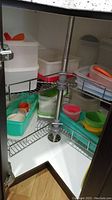 Food storage containers and colorful lids stacked on two rotating wire shelves inside a corner kitchen cupboard.