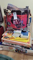 Stack of vintage board games on a carpeted floor, with a heavily damaged box containing a play tent on top, along with a pile of books and VHS movies.
