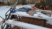 Garden tools with wooden handles including shovels, rakes, saw, clippers, and weed puller placed on a picnic table outside with snow ground.