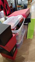 Stack of eight black and red storage boxes next to two clear plastic bins with decorations inside and a green wooden shelf leaning against them.
