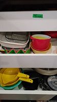 Two shelves showing nested bowls in red, yellow, and white, green and yellow plastic strainers, and assorted kitchenware items including a black pan and containers.