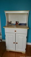Front view of microwave cart showing two doors, one drawer and open shelf at the top with various small items on it.