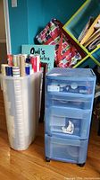 Wrapping paper rolls in white plastic bin and blue plastic 4-drawer storage unit to the right