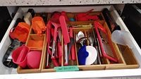 Drawer of assorted kitchen utensils organized in wooden tray with multiple compartments. Various items in red, orange, black, and metal.