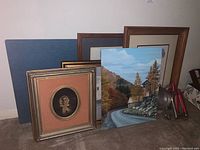 Group of framed artworks including vintage photo and signed prints beside acrylic painting and three metal candleholders on floor against wall.