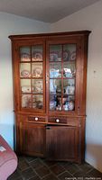 Full view of wooden corner cabinet with glass doors displaying dishware inside, two drawers below, and two wooden lower doors closed.