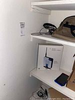 Photo of shelves showing the Linksys wireless router box labeled T-Mobile HotSpot Home, headphones, tan shoulder bag, and a Samsung phone with blue case.