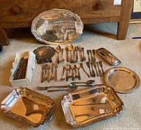 Photo showing multiple silver-plated serving trays, flatware pieces arranged on carpeted floor, and a decorative trivet. Items appear tarnished and used.
