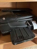 Photo showing black HP Officejet 6700 Premium printer with some dust, ink cartridge installed, keyboard behind printer.
