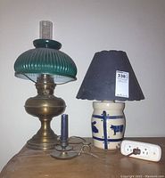 Photo showing the brass lantern lamp with green ribbed glass top, ceramic country cow pattern lamp with dark scalloped fabric shade, small brass candle holder with candle and music stand style handle, and white three plug adapter on the table.