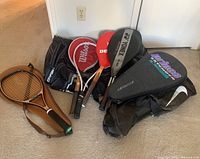 Photo showing five racquets (two Dunlop with red covers, one Wilson with red cover, and two Yonex; one with grey cover) resting on two black sports bags, one Nike branded and one Tour Team.