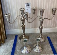 Four sterling silver candleholders displayed on carpet in front of wall. Two are larger three-arm candelabra, two are smaller single holders. Shows overall tarnish.