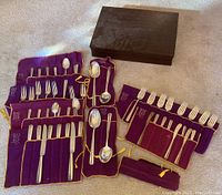 Wooden tarnish-resistant chest alongside purple cloth rolls holding multiple flatware pieces and loose serving spoons.