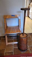 Photo showing wooden folding chair and vintage butter churn placed side by side on tiled floor
