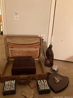 Overview photo showing open vintage Hartmann leather briefcase with several interior pockets, a dark wood box inside the briefcase, two humorous plaques, a piece of rock, a wooden carved eagle, and a silver-tone ashtray on carpeted floor.