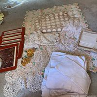 Overview of all linens including crocheted baby blanket, tablecloth, doilies, stacked cloth napkins and handkerchiefs along with framed hot pads.