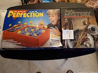 Photo showing Perfection board game box with pieces, and Murder Mystery Party game box side by side.