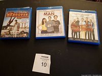 Photo of three Blu-ray DVDs on black surface showing The Hangover (Unrated), I Love You Man, and The Other Guys with a MaxSold numbered tag.