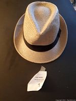 Front side view of the beige straw fedora hat with black band, showing the pinched crown and attached tag below.