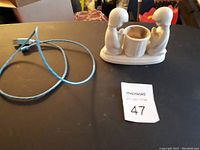 Photo showing ceramic candle holder with two abstract figures kneeling, holding a circular candle cup, and a blue charging wire with plug laying beside it.