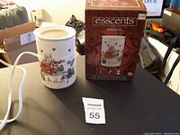 Photo showing white ceramic electric candle warmer holder decorated with red cabin, snowman, evergreen trees and reindeer in a snowy landscape along with original box and lot number.
