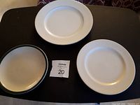 Photo shows three different plates on a dark surface: one plain white with smooth rim, one white with textured/embossed rim, and one cream-colored with dark rim.