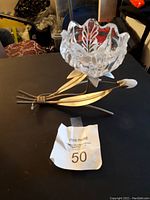 Photo showing a metal flower-shaped candle holder with a clear glass cup insert to hold a candle, placed on a dark surface with a MaxSold tag.
