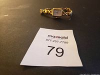 Small gold-tone women's wristwatch with linked decorative band, square white dial with black numbers and gold-tone accents, photographed on black surface with auction lot number paper.