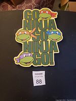 Flat image of colorful Teenage Mutant Ninja Turtles hanging sign with text.