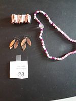 Photo showing a pink beaded necklace with a central clear bead, a pair of gold-tone leaf-shaped dangle earrings, and a beige and brown striped clip-style bracelet on a black background.