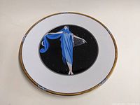 Full view of the white charger plate with a central black circle featuring a blue robed figure and gold trimmed edges.