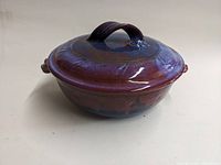 Angled side view of large covered casserole showing purple and blue glaze, lid with twisted handle, and side handles.