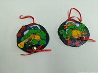 Pair of Vintage Teenage Mutant Ninja Turtles Christmas Ornaments, showing both ornaments with red ribbons and bells.