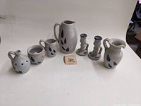 Full view of seven pieces of grey glazed Williamsburg stoneware pottery with blue leaf and berry decorations arranged in a row on a white surface.