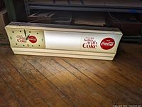 Full view of the rectangular lighted Coca-Cola sign with clock face printed on left side and Coca-Cola logo with slogan on right side, placed on wooden floor.