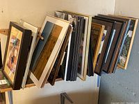 Stack of various framed art pieces and photos leaning against a wall on a table, showing diverse frame styles and sizes.
