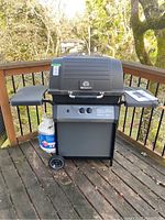 Front view of black BroilMate gas barbecue grill on wooden deck, with propane tank attached beneath on left side, and two side tables.