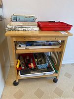 Full view of the wooden kitchen cart on wheels with trays of utensils on shelves and cookbooks on top.