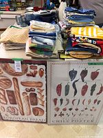 Kitchen linens displayed folded in stacks showing various colors and patterns including blues, yellows, whites, and reds. Two framed posters at front depicting European bread and chili peppers.