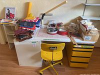 Desk assembly showing tabletop, small shelf, dresser, yellow cabinet and chair with fabrics piled on top
